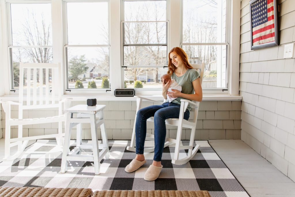 A woman sitting in a white rocking chair, holding her newborn with an American flag on the wall behind her.